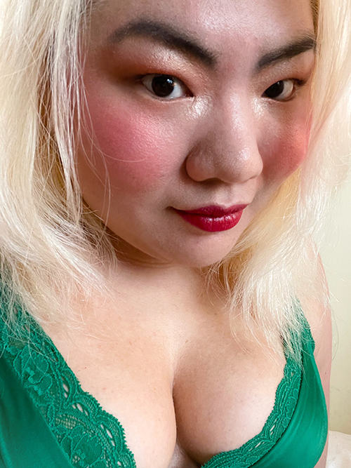 Amber Ying: A blonde feminine person is in three-quarter view smiling slightly at the viewer. They are wearing makeup: rust orange eyeshadow, pink blush, and red lipstick. They are also dressed in a green tanktop with lace trimming.
